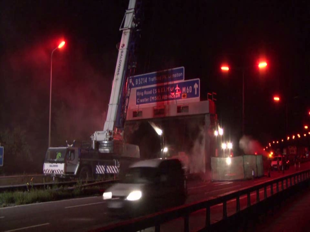 A motorway at night. Near the camera a small van is passing. Further away, beyond a barrier, a large crane on the back of a stationary truck stretches up into the darkness, as it prepares to remove a gantry that carries motorway signs.
